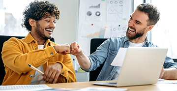 Two men in an office fist-bumping and celebrating their success.