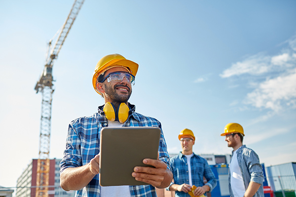 A construction worker is holding a tablet and smiling at a job site.