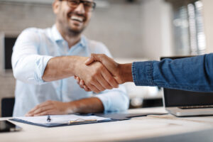 Two people shaking hands with one man in the background smiling.